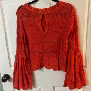May include: Bright orange long-sleeved top with bell sleeves. The top features a keyhole neckline and intricate open-knit detailing. The sleeves have a flared design, and the hemline is scalloped. The top is hanging on a black hanger against a white door.