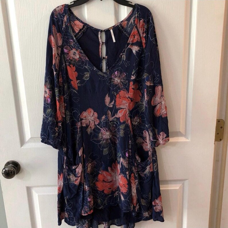 K&ouml;nnte beinhalten: Ein marineblaues Kleid mit einem Blumenmuster in Rot-, Rosa- und Wei&szlig;t&ouml;nen. Das Kleid hat lange &Auml;rmel, einen V-Ausschnitt und zwei Vordertaschen. Das Kleid h&auml;ngt an einem schwarzen Kleiderb&uuml;gel.