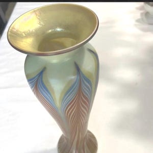 Può includere: Un vaso decorativo in vetro con una parte superiore svasata e un corpo sottile e curvo. Il vaso presenta un colore di base verde pallido con motivi verticali a forma di piuma in blu, marrone e giallo. Il bordo ha una finitura dorata.