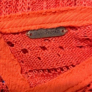 May include: Close-up of a coral-colored knit top with a textured design. The top features a small, rectangular metal tag with the words "Free People" in a cursive font. The fabric has a slightly open weave.
