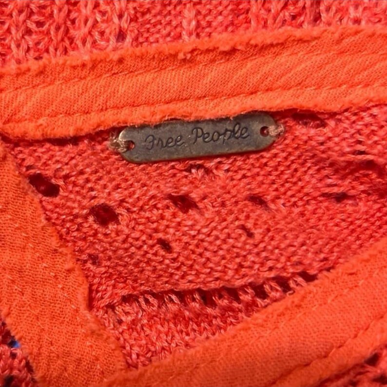 May include: Close-up of a coral-colored knit top with a textured design. The top features a small, rectangular metal tag with the words "Free People" in a cursive font. The fabric has a slightly open weave.