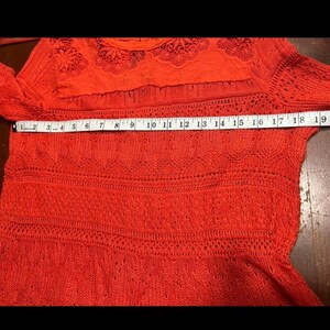 May include: A bright orange sweater with a lace yoke and long sleeves. The sweater has a textured knit design with a measuring tape laid across the front, showing a measurement of approximately 19 inches.