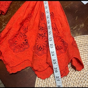 May include: Bright orange embroidered top with a scalloped hem. The top features intricate floral cutouts and is measured with a tape measure. The length of the top is approximately 27 inches.