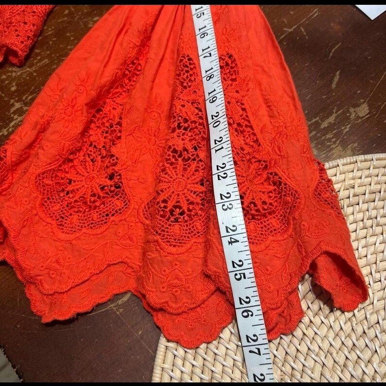 May include: Bright orange embroidered top with a scalloped hem. The top features intricate floral cutouts and is measured with a tape measure. The length of the top is approximately 27 inches.