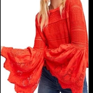 May include: A bright red long-sleeved top with flared sleeves. The top features intricate lace detailing on the sleeves and bodice. The sleeves have a scalloped edge. The top is paired with blue jeans.