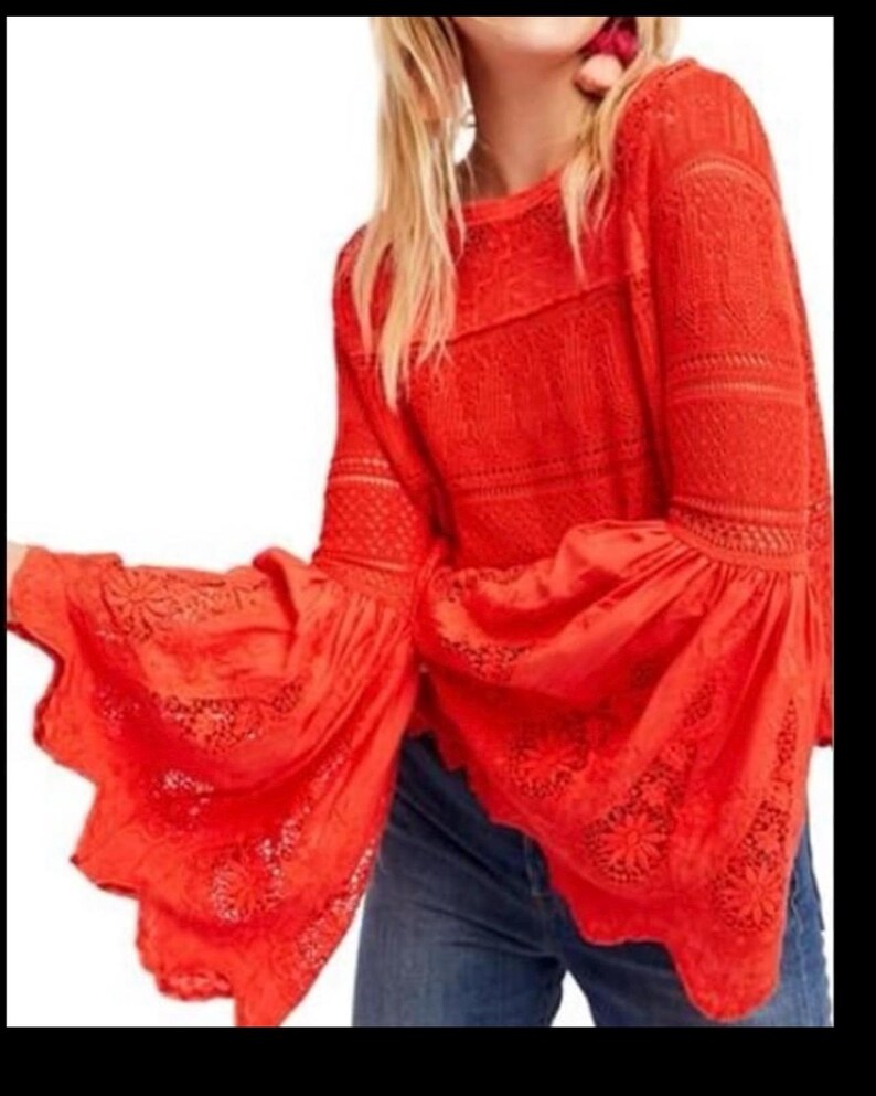 May include: A bright red long-sleeved top with flared sleeves. The top features intricate lace detailing on the sleeves and bodice. The sleeves have a scalloped edge. The top is paired with blue jeans.