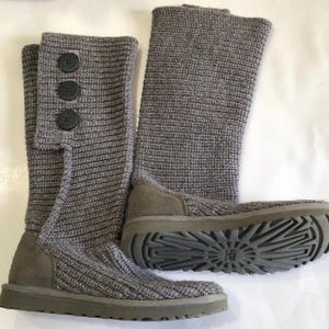 May include: A pair of gray knit boots with a button closure on the side. The boots have a suede heel and sole, and the soles are a darker gray color. The boots are made of a thick knit material.