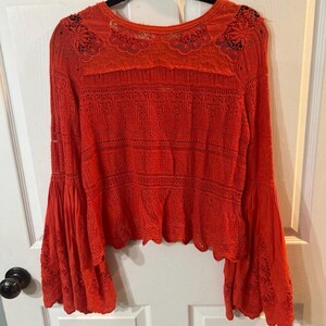 May include: Bright red long-sleeved top with bell sleeves. The top features intricate lace detailing across the chest, shoulders, and sleeves. The hemline has a scalloped edge. The top is made of a lightweight, possibly woven material.