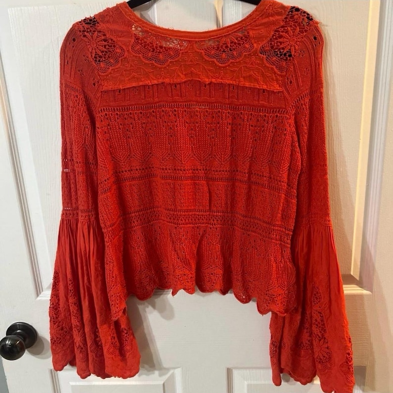 May include: Bright red long-sleeved top with bell sleeves. The top features intricate lace detailing across the chest, shoulders, and sleeves. The hemline has a scalloped edge. The top is made of a lightweight, possibly woven material.