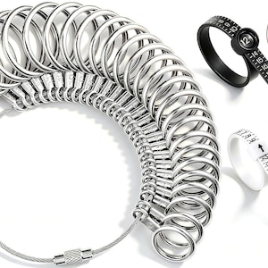 May include: A set of silver-colored metal ring sizers arranged on a circular wire cable, accompanied by one black plastic ring sizer and one white plastic ring sizer. The black sizer displays the number 12 in a circular window with markings.