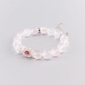 Könnte beinhalten: Armband aus Rosenquarzperlen mit einem silbernen Mittelstück. Das Mittelstück hat einen ovalen rosa Edelstein, der von einer dekorativen silbernen Fassung umgeben ist. Das Armband hat einen zarten Kettenverschluss.