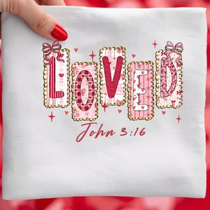 May include: White fabric with the word "LOVED" in pink, red, and gold letters. Each letter is decorated with hearts, bows, and a scalloped border. The text "John 3:16" is written in red script below the word "LOVED".