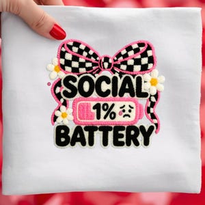 Puede incluir: Tela blanca con un diseño bordado colorido. El diseño incluye un lazo a cuadros blanco y negro, las palabras "SOCIAL BATTERY", un icono de batería que muestra el 1% y pequeñas flores de margarita.