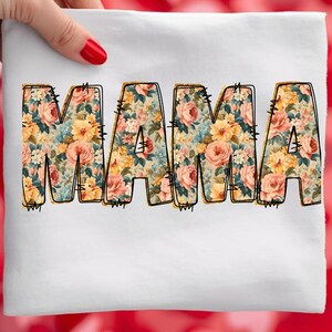 Könnte beinhalten: Ein weißes Stoffstück mit dem Wort "MAMA" in großen Blockbuchstaben. Die Buchstaben sind mit einem Blumenmuster in Rosa, Gelb und Blau gefüllt. Die Buchstaben sind schwarz umrandet und mit dekorativen Nähten versehen.