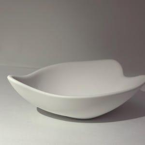 Op de afbeelding: Een witte, bladvormige keramische kom. De kom heeft een gladde, matte afwerking en zacht gebogen randen. Het ontwerp is minimalistisch en modern, geschikt om te serveren of als decoratief stuk. De kom is leeg en staat op een wit oppervlak.