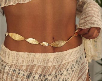 May include: A gold-colored waist chain with leaf-shaped links. The chain is worn over a cream-colored lace outfit. The leaves are textured and connected by small links. The overall style is bohemian and elegant.