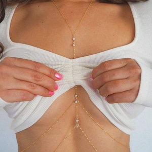 Op de afbeelding: Een gouden lichaamsketting met meerdere strengen en kleine, heldere edelstenen. De ketting is ontworpen om over de borst en torso te draperen. De sieraden worden gedragen over een witte top met lange mouwen.