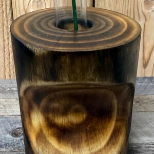 May include: A handcrafted wooden vase with a clear glass insert for a single stem flower. The vase is made from a dark, stained wood, showcasing the natural grain and circular growth rings. The vase is cylindrical and has a rustic, natural aesthetic.