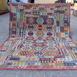 Könnte beinhalten: Ein großer, rechteckiger Teppich mit einem lebendigen geometrischen Muster. Der Teppich weist eine Vielzahl von Farben auf, darunter Rot, Blau, Orange, Gelb und Grün. Das Design umfasst Diamanten, Sechsecke und rechteckige Formen. Der Teppich wird im Freien ausgestellt.
