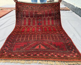 May include: A rectangular area rug featuring a deep red base color with intricate black and dark brown geometric patterns. The design includes repeating arch motifs and a central vertical column, finished with light-colored fringe at the bottom edge.
