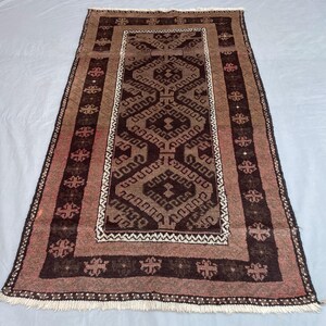 Pode incluir: Um tapete retangular, tecido à mão, com um desenho geométrico central em tons de castanho e bege. O tapete apresenta uma borda com motivos florais repetidos e franjas nas bordas. Dimensões: aproximadamente 91 x 152 cm.