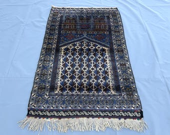 May include: A rectangular prayer rug featuring a deep blue and dark brown color palette with a central mihrab arch design. The rug displays intricate geometric patterns, a detailed border, and white fringe along the bottom edge of the textile.