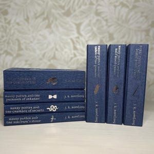 May include: A collection of Harry Potter books in a navy blue color. The titles and author's name, J.K. Rowling, are printed in silver. The books are stacked and arranged, showcasing the series' titles and iconic symbols.