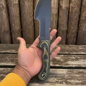 May include: A black and silver knife with a curved blade and a serrated edge. The handle is green and brown with three silver rivets. The knife is held in a hand against a wooden background.