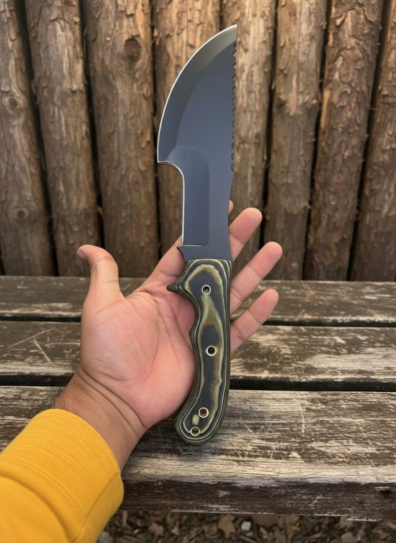 May include: A black and silver knife with a curved blade and a serrated edge. The handle is green and brown with three silver rivets. The knife is held in a hand against a wooden background.