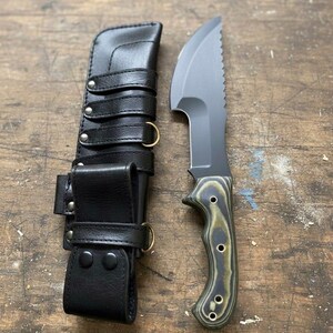 May include: A black knife with a serrated edge and a green and black handle, next to a black leather sheath with straps and metal rings. The knife blade is gray. The handle has a wood grain pattern. The sheath has multiple straps and metal hardware.