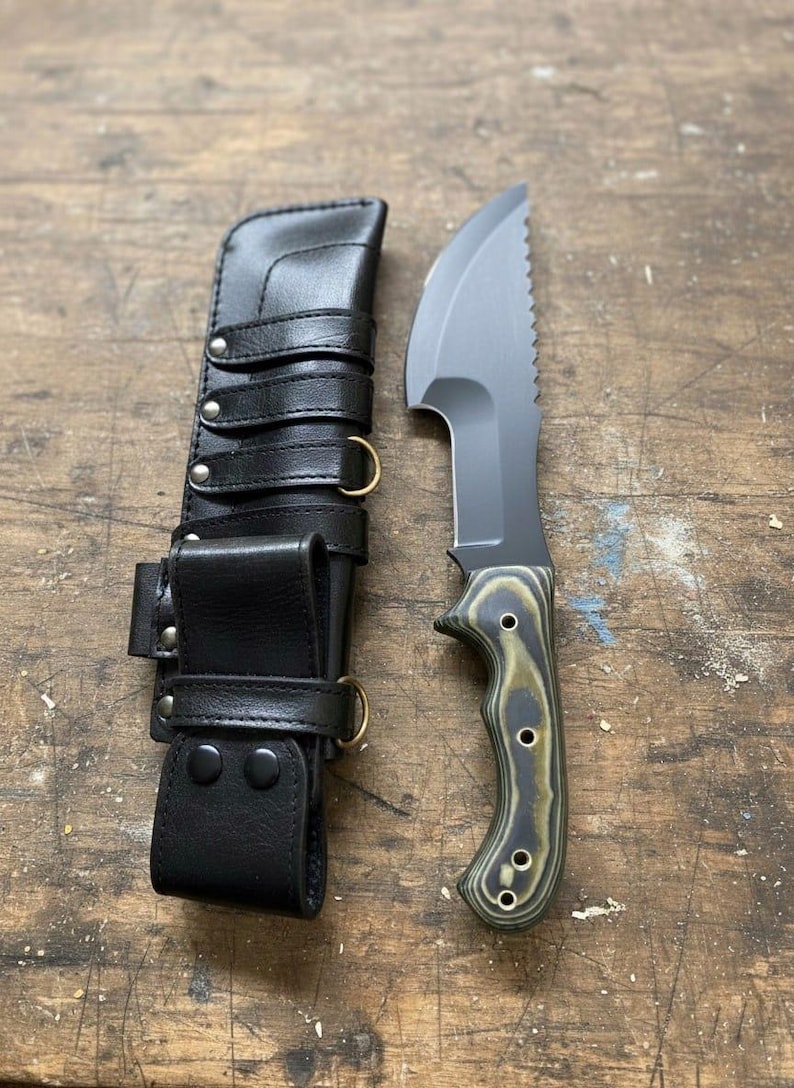 May include: A black knife with a serrated edge and a green and black handle, next to a black leather sheath with straps and metal rings. The knife blade is gray. The handle has a wood grain pattern. The sheath has multiple straps and metal hardware.