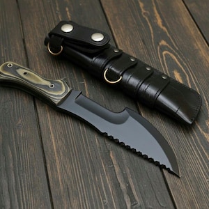 May include: A black hunting knife with a serrated edge and a wooden handle, next to a black leather sheath. The knife has a curved blade and a dark finish. The sheath has a loop and snap closure.