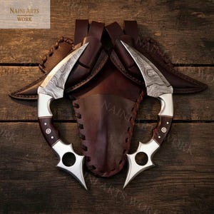 May include: Two curved knives with patterned blades and dark wood handles, resting on a brown leather sheath. The handles have silver accents and decorative rivets. The image includes the text "NAINI ARTS WORK".