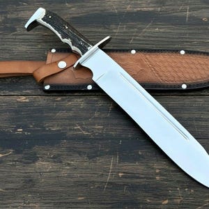 May include: A large, polished steel knife with a black and white handle rests on a brown leather sheath. The knife has a wide blade and is designed for outdoor use. The sheath is secured with a leather strap and a metal stud.