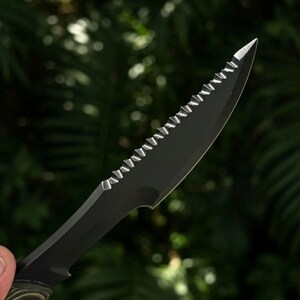 May include: Close-up of a black serrated knife with a pointed tip. The blade has a series of sharp teeth along the top edge. The handle is a mix of green and brown tones. The background is a blurred view of green foliage.