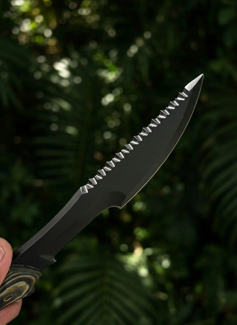 May include: Close-up of a black serrated knife with a pointed tip. The blade has a series of sharp teeth along the top edge. The handle is a mix of green and brown tones. The background is a blurred view of green foliage.