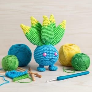 May include: A crocheted blue character with green and yellow accents, pink eyes, and small blue feet. The character is surrounded by balls of yarn in teal, green, and yellow, along with knitting needles and a crochet hook. The theme is handmade crafts.