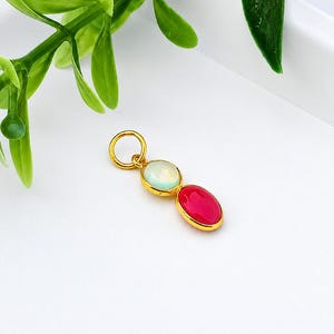 Puede incluir: Colgante dorado con dos piedras preciosas de colores: un óvalo azul claro y un óvalo rojo. El colgante está sobre una superficie blanca con hojas verdes en el fondo.