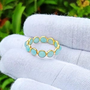 Könnte beinhalten: Ein goldener Ring mit einer durchgehenden Reihe kleiner, runder, hellblauer Cabochon-Steine, die in einzelnen Goldfassungen gefasst sind. Der Ring wird vor einem unscharfen grünen Hintergrund präsentiert und zeigt das polierte Goldmetall sowie die glatte, opake Textur der kreisförmig angeordneten hellblauen Steine.