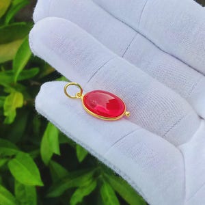 May include: A gold pendant featuring a smooth, oval-shaped, translucent red gemstone set in a polished gold bezel. The pendant includes a small, circular gold jump ring at the top for attachment, showcasing a vibrant red hue and metallic gold finish.