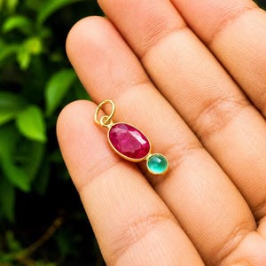 May include: A gold pendant featuring a large oval faceted deep pink gemstone and a smaller round smooth light green gemstone set in a bezel setting with a circular jump ring attached at the top for hanging on a chain or cord.