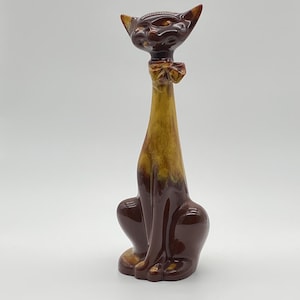 May include: A tall, stylized ceramic cat figurine featuring a glossy finish with a gradient color scheme. The base and head are a deep, dark brown, while the elongated neck and chest area transition into a lighter, mottled golden-brown hue.