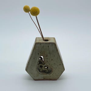 以下が含まれることがあります： 六角形で斑点のある仕上げの小さなオフホワイトの陶器製花瓶。花瓶の中央には切り込みがあり、内部の渦巻き状の粘土のディテールが見えます。上部の開口部には、2本の乾燥した丸い黄色の花が挿されています。