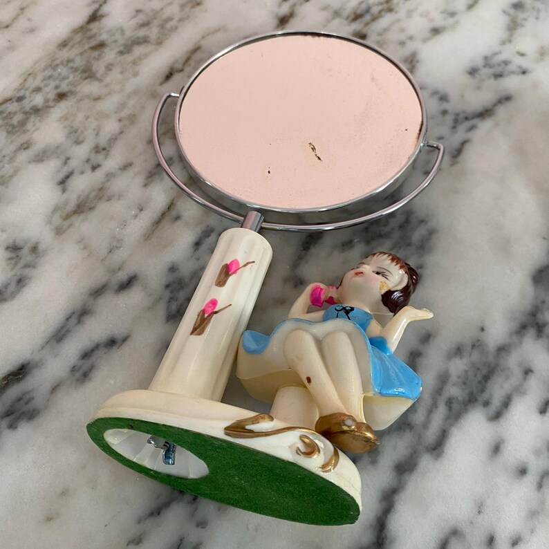 May include: A vintage vanity mirror with a round, light pink mirror and a decorative base. The base features a seated figurine in a blue dress and gold shoes. The mirror's handle is cream-colored with pink floral accents.