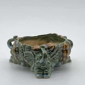 May include: A ceramic planter featuring three sculpted elephant heads integrated into the base and sides. The piece has a glossy, multi-toned glaze in shades of sage green, olive, and brown, with a smooth, light-colored interior basin.