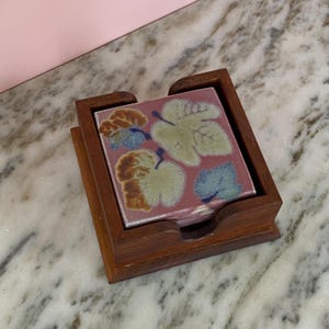 Peut inclure: Un ensemble de quatre dessous de verre carrés en céramique avec un motif de feuilles dans un support en bois. Les dessous de verre ont un fond rose avec des motifs de feuilles verts, bleus et marron. Le support en bois est de couleur marron foncé.