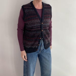 May include: A patterned sweater vest in navy, burgundy, and grey, with a V-neck and wooden buttons. It is worn over a long-sleeved burgundy top and paired with blue jeans. The vest's pattern is a mix of textures.