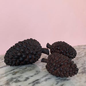 May include: Three dark brown decorative pine cones with textured surfaces. The pine cones are arranged on a marble surface against a pink background. The pine cones are a rustic home decor item.