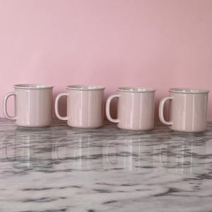 May include: Four pale pink ceramic mugs with white rims and handles are arranged in a row. The mugs are on a marble surface, with a solid pink background. Each mug is the same shape and size.