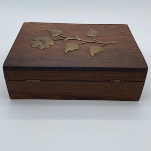Peut inclure: Une boîte rectangulaire en bois avec une finition à grain brun foncé, ornée d'une incrustation en laiton représentant une fleur et des feuilles sur le couvercle. La boîte possède un couvercle à charnières avec deux charnières en laiton visibles.